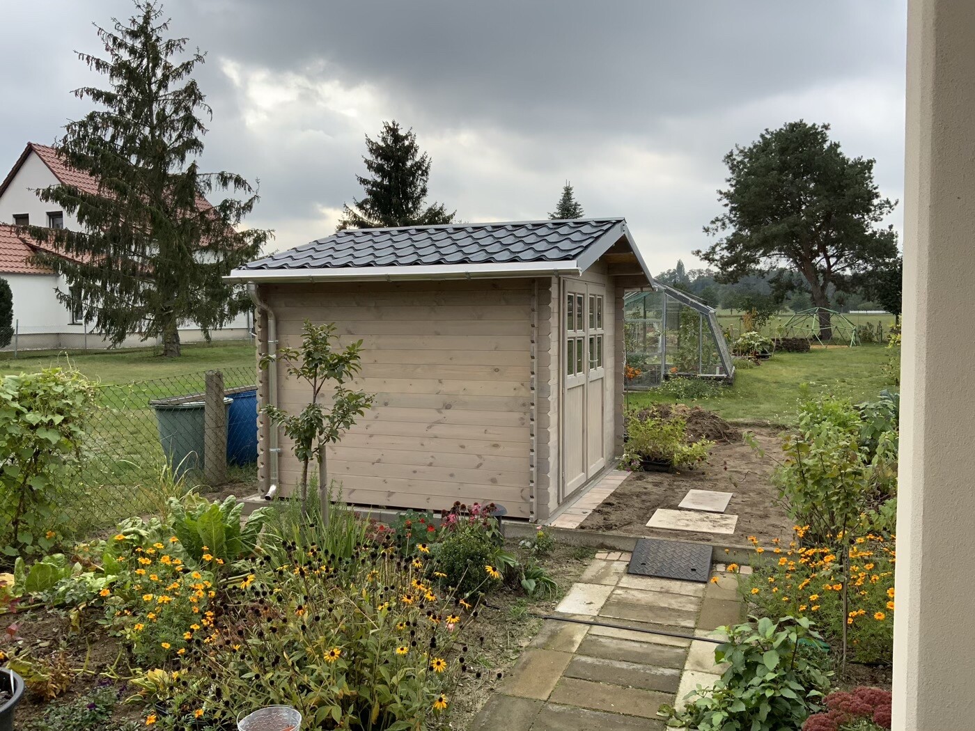 Kleines Holz-Gartenhäuschen mit dunklem Dach, Blumenbeeten und einem Gewächshaus im Hintergrund.