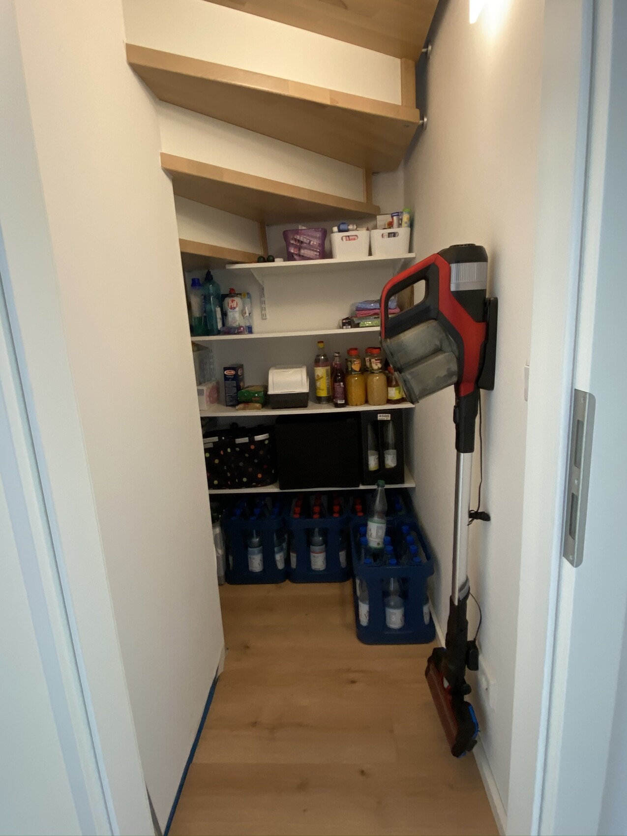 Kleiner Abstellraum unter der Treppe mit Regalen voller Reinigungsmittel, Getränke und Staubsauger.