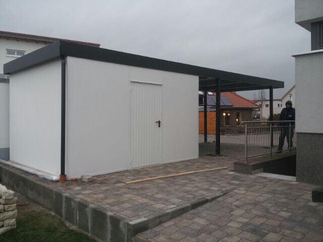 Kleiner weißer Schuppen mit schwarzem Dach auf gepflastertem Vorplatz neben Wohnhäusern.