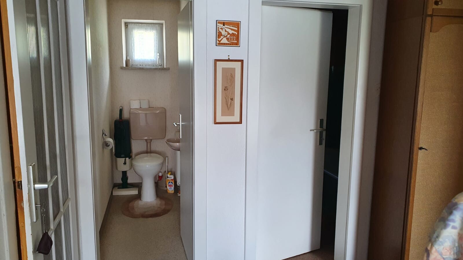 Kleiner Flur mit offener Badezimmertür: Toilette, Waschbecken, weißer Schrank und Bilder.