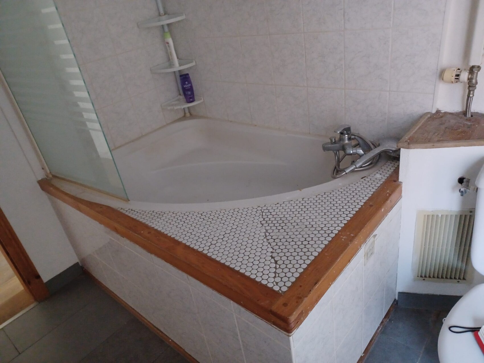 Eckbadewanne mit Glasabtrennung, Mosaikrand aus weißen Fliesen, Holzrand, Duscharmatur und Shampoo.