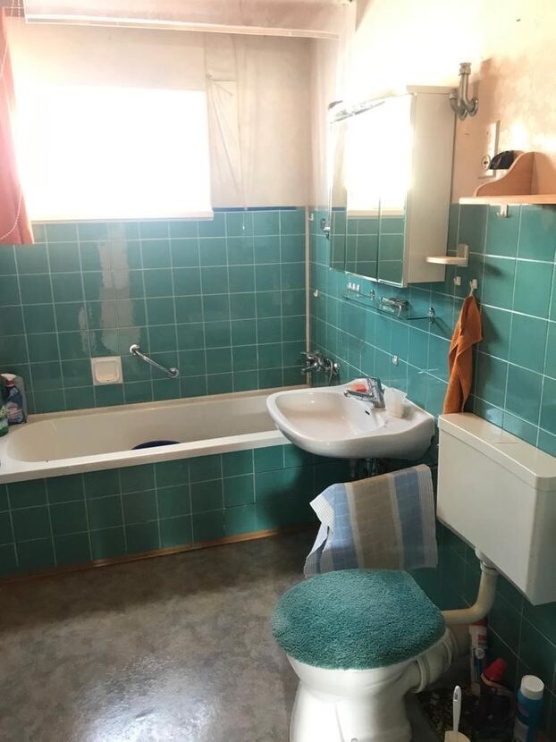 Kleines Bad mit grünen Fliesen, Badewanne links, Waschbecken, Toilette mit blauem Deckel, Handtuch.