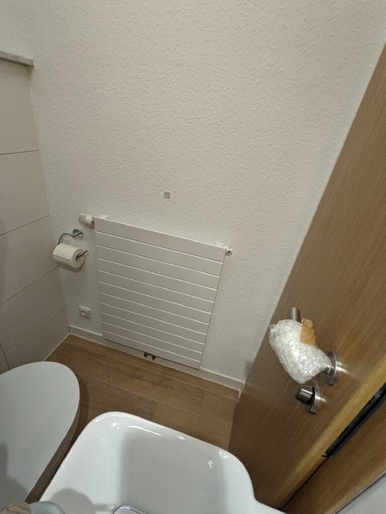 Kleines Badezimmer mit Heizkörper, Toilette, Toilettenpapierhalter und offener Tür.