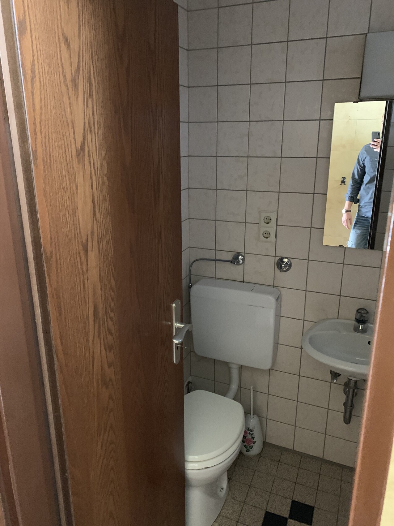 Kleines Bad mit Toilette, Spülkasten, Waschbecken; Spiegelbild einer Person.