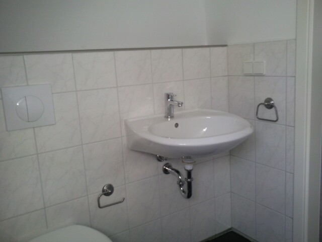 Kleines Badezimmer mit wandhängendem Waschbecken, sichtbaren Rohren und Fliesen.