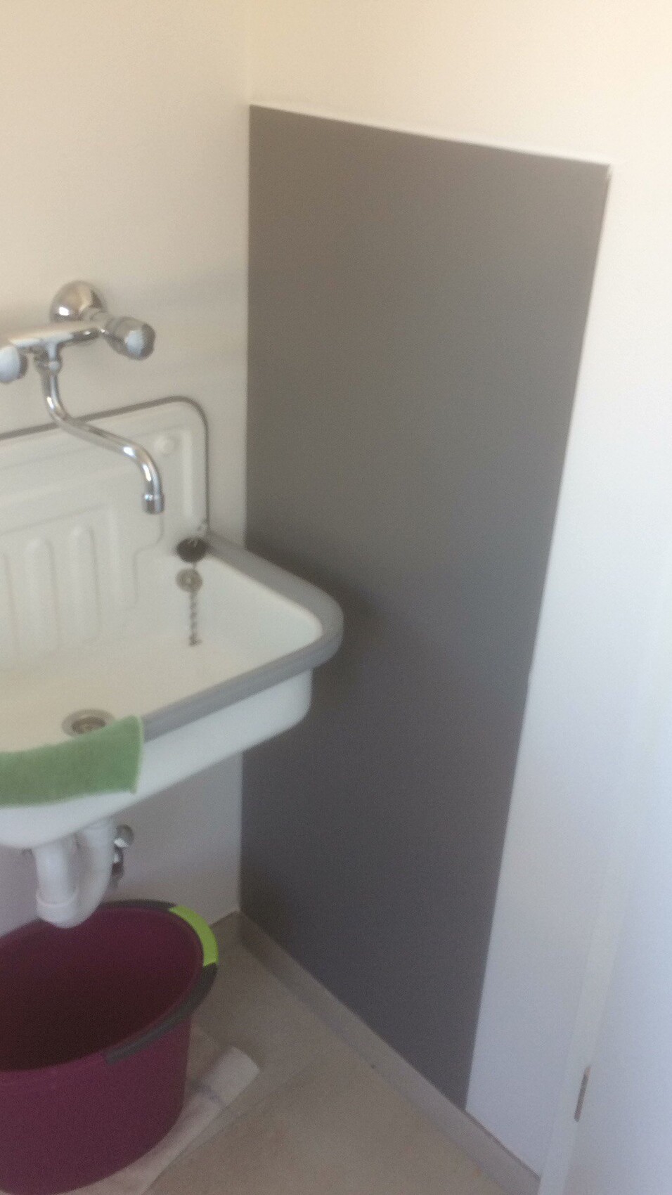 Kleines Waschbecken mit Metallarmatur, graues Wandpaneel und pinker Eimer darunter.