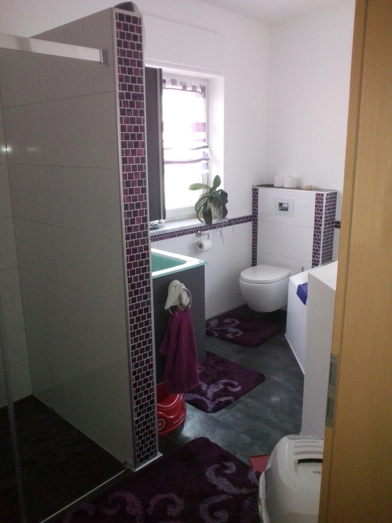 Badezimmer mit violetten Mosaikfliesen, Dusche, Wandtoilette, Fensterpflanze und Teppiche.
