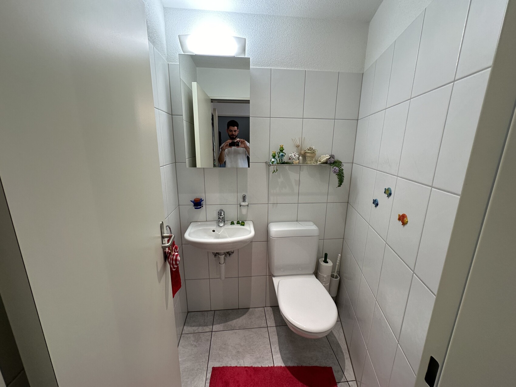 Kleines Badezimmer: Waschbecken, Toilette, Spiegel; Mann fotografiert sich im Spiegel.