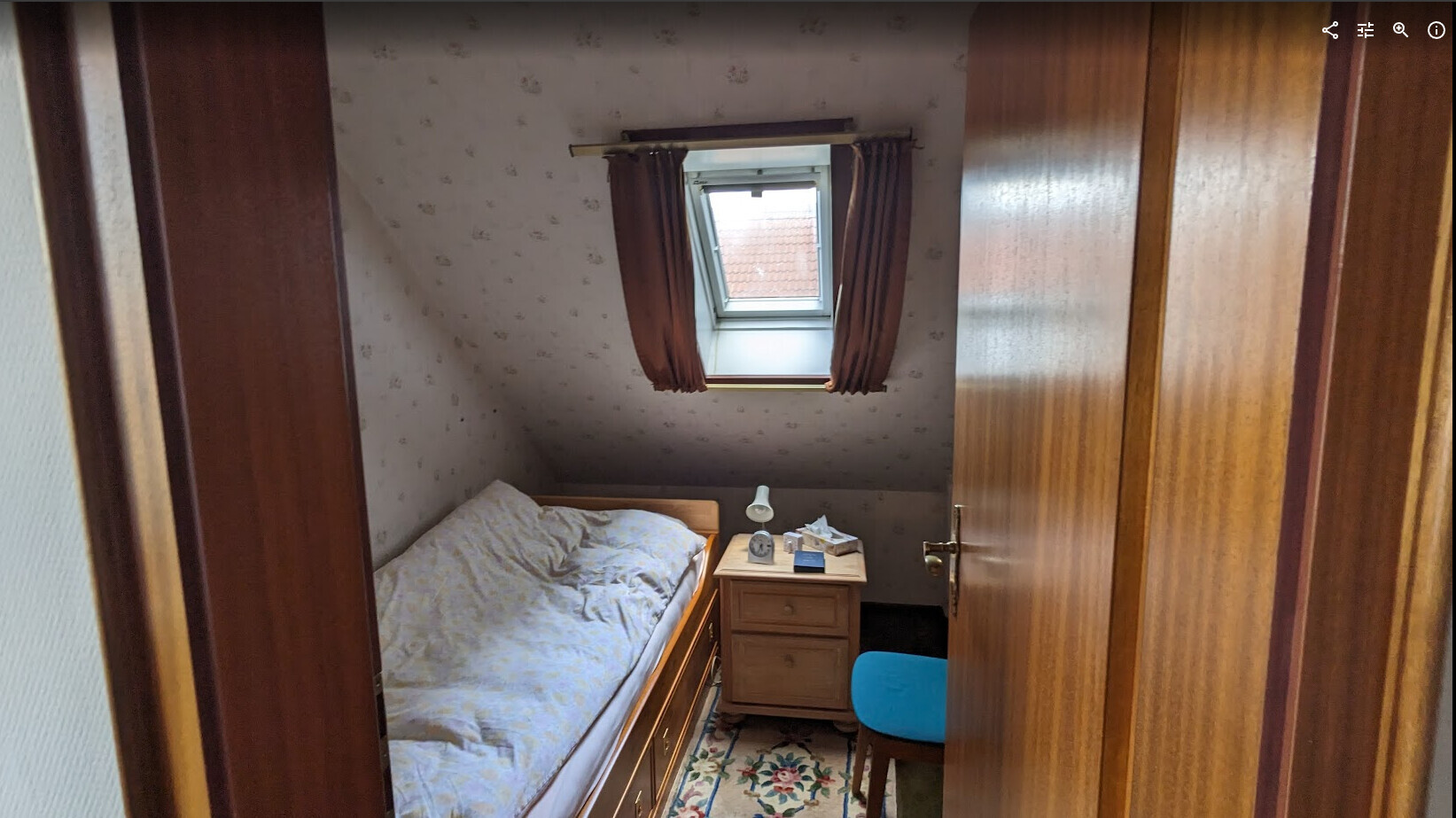 Kleines Schlafzimmer mit Dachfenster, Bett, Nachttisch, Lampe und blauem Stuhl.