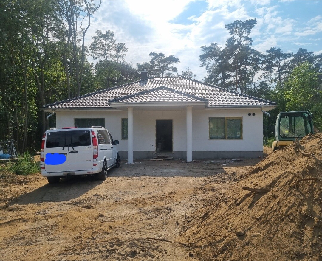 Weißes Einfamilienhaus mit Ziegel-Dach, zwei Säulen am Eingang; links Van, rechts Bagger.