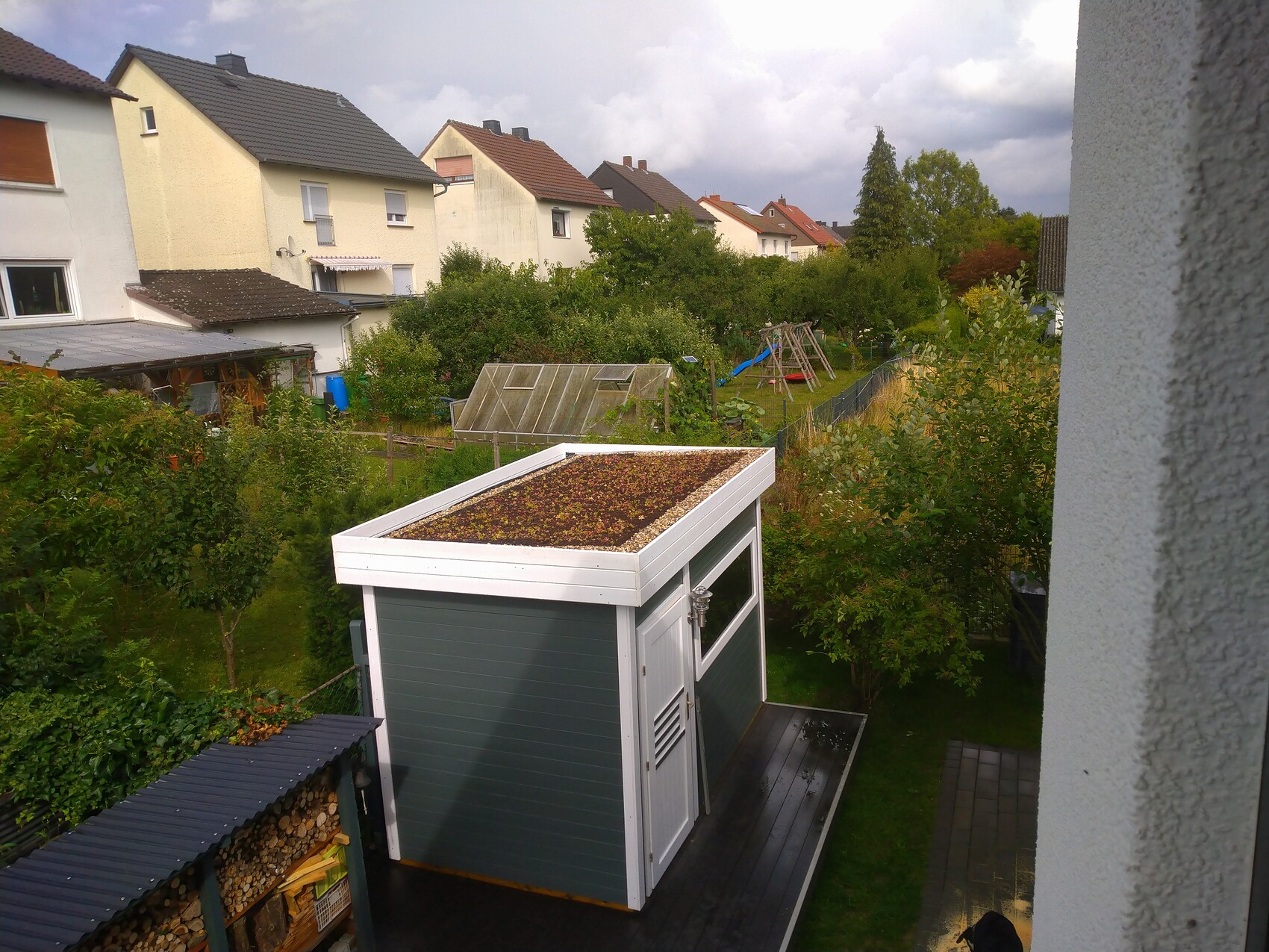 Kleiner Garten mit weiß-grünem Gartenhaus, Moos auf dem Flachdach, Spielplatz im Hintergrund.