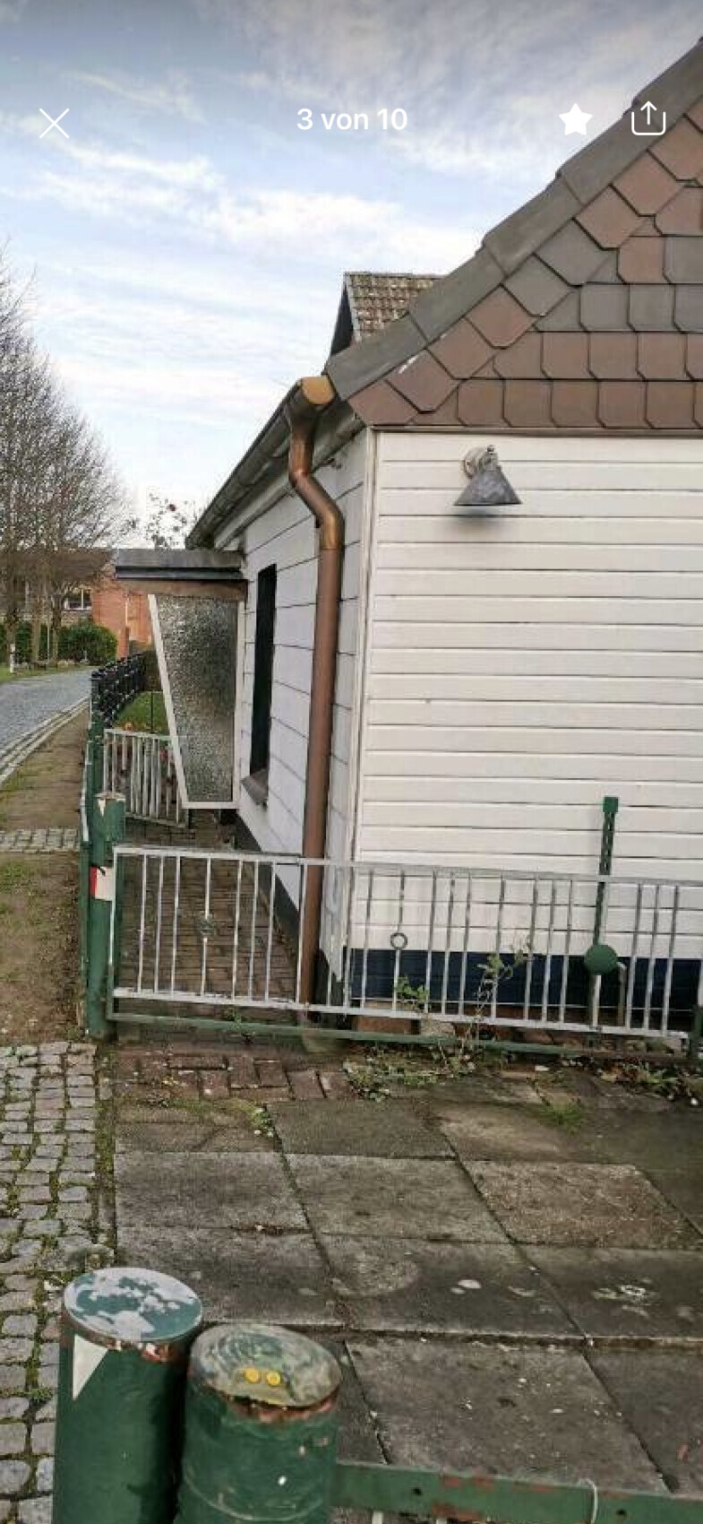 Vorderseite eines kleinen weißen Holzhauses mit braunem Dach, Kupfer-Rinne, Zaun und grünen Tonnen.