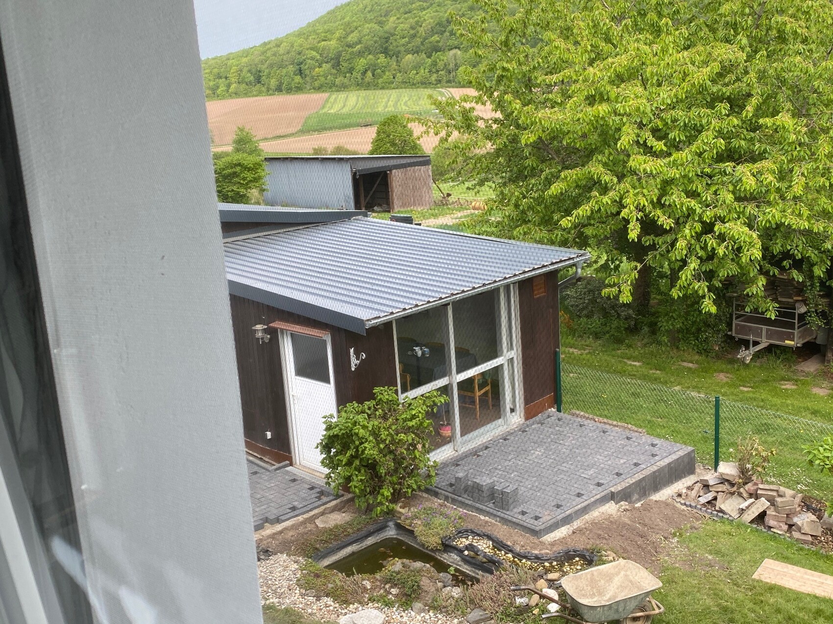 Gartenhaus mit Metall-Dach, Glasfront und Steinplatten-Terrasse; Baum, Garten und Felder dahinter.