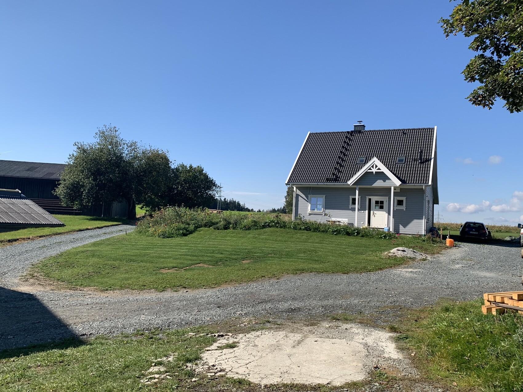 Kleines graues Haus mit schwarzem Dach, Garten, Kiesweg und Auto am Straßenrand vor blauem Himmel.