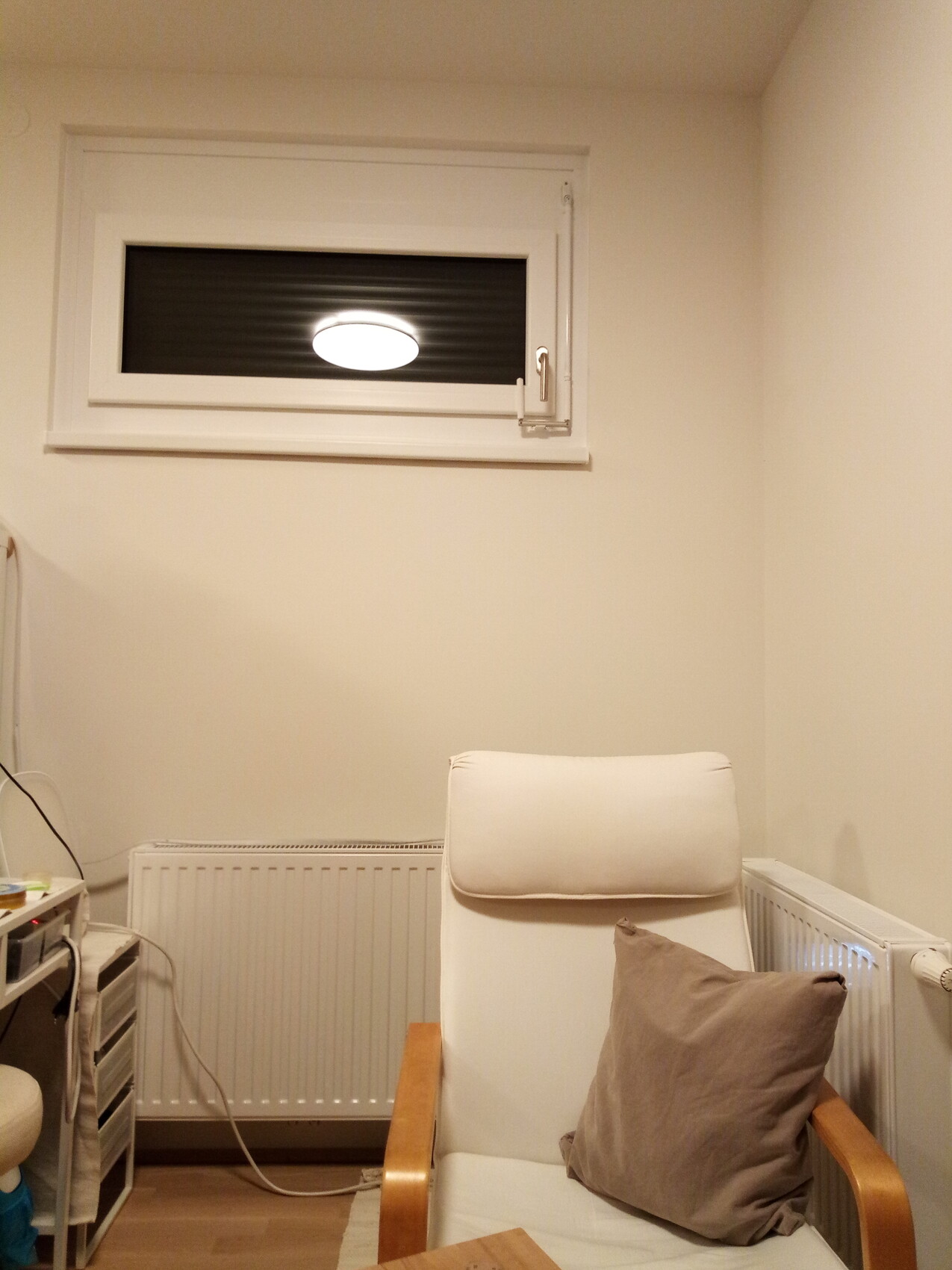 Weißer Lehnstuhl mit braunem Kissen vor Heizkörper und Fenster hoch an der Wand.