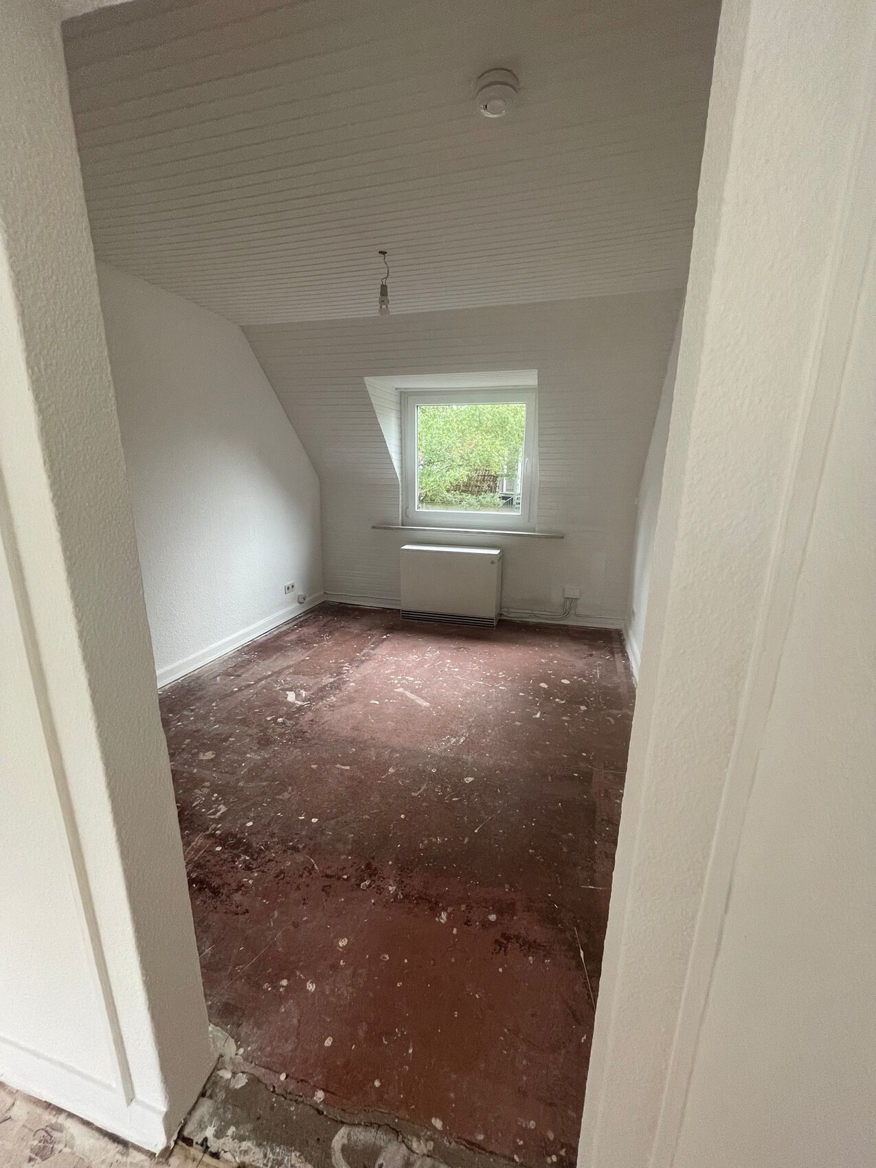 Kleines Dachzimmer mit schräger Decke, Fenster zur Grünfläche, Heizkörper, roher Boden, leer.