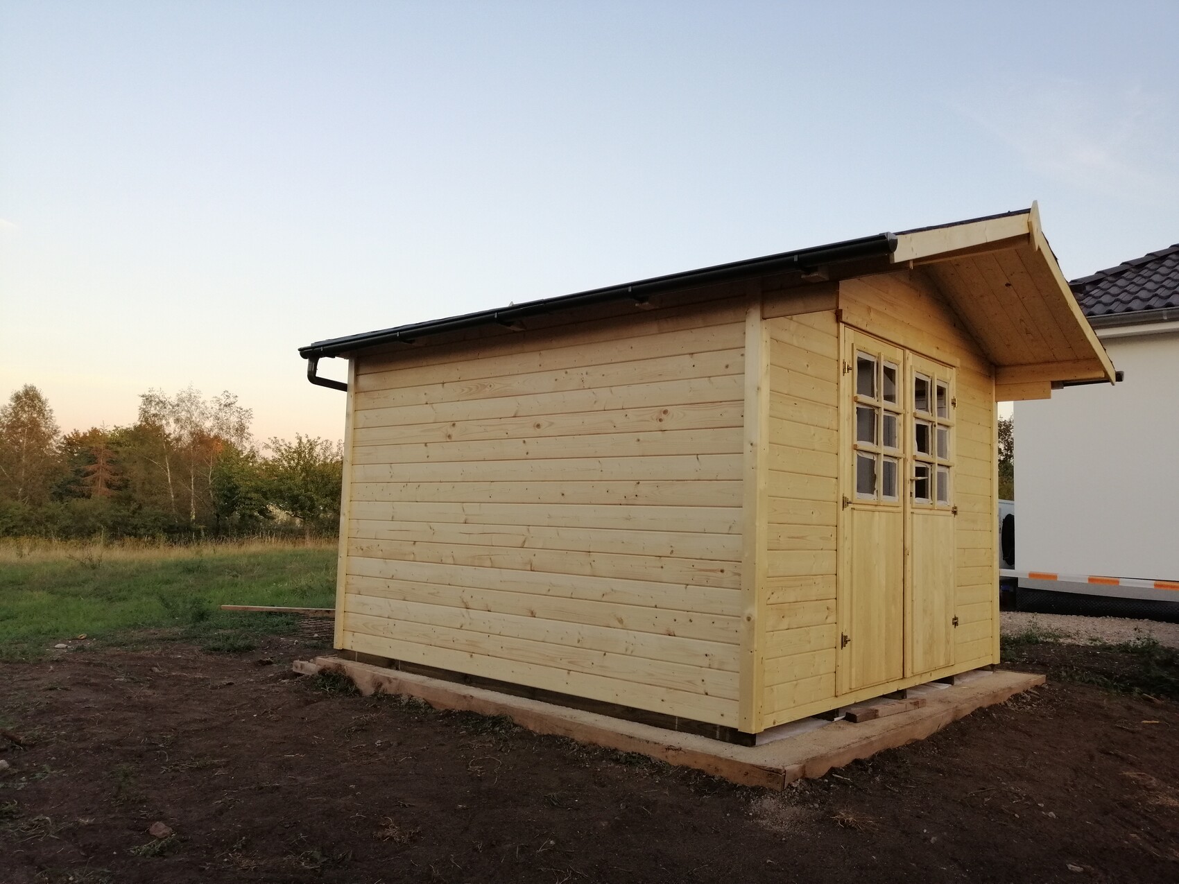 Kleiner Holzschuppen aus Naturholz mit Tür und Fensterfront, auf Holzfundament im Freien.
