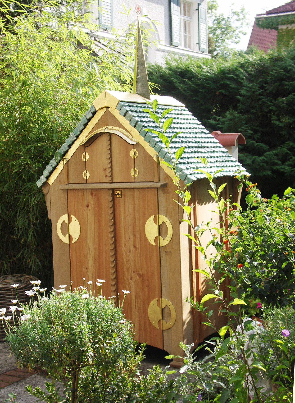Kleines Holzgartenhaus mit grünem Ziegeldach, goldenen Ringen, von Pflanzen umgeben.
