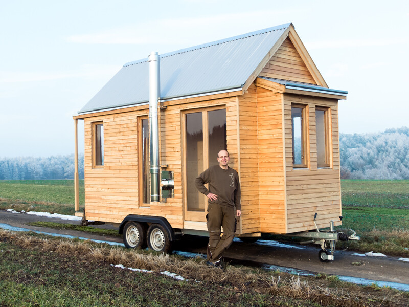 Kleines Holzhaus auf einem Anhänger, Mann steht davor, ländliche Landschaft.