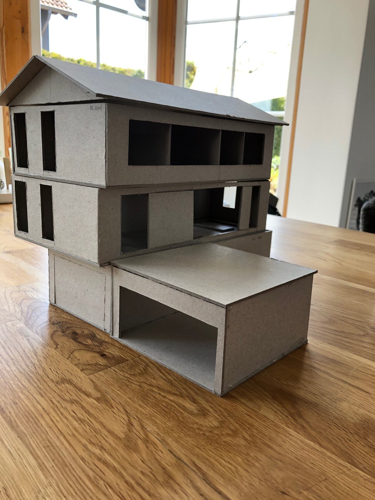 Kartonmodell eines zweistöckigen Hauses mit offenen Fensteröffnungen auf Holztisch.