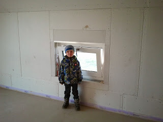 Kleines Kind in Winterkleidung mit Helm steht auf unfertiger Innenbaustelle vor einer Fensteröffnung