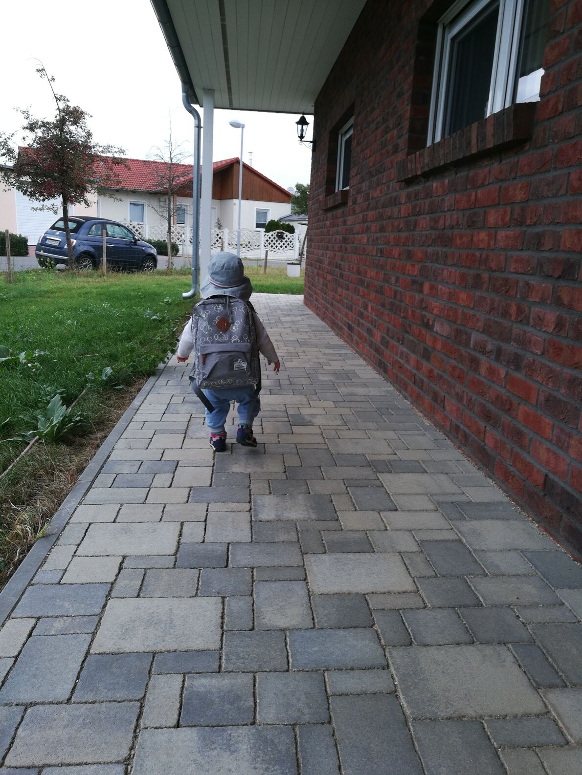 Kleines Kind mit grauem Rucksack läuft auf gepflastertem Weg neben rotem Backsteinhaus.