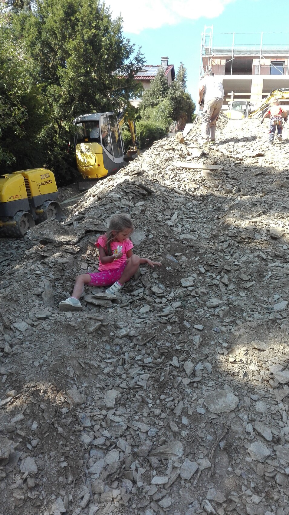 Kleines Mädchen in Pink sitzt auf einem Kieshügel auf Baustelle; Baumaschinen im Hintergrund