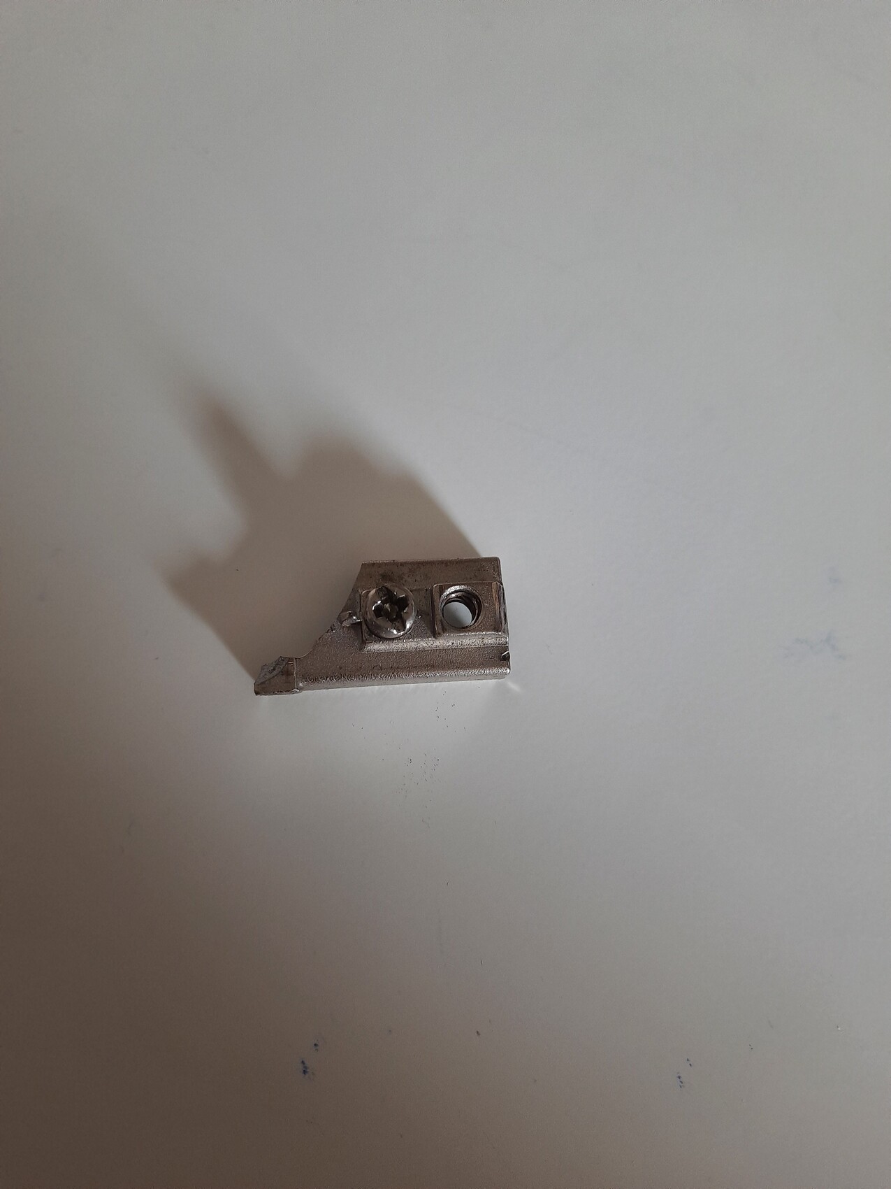 Kleines metallisches Montagesteil mit Schraube und zwei Bohrungen auf heller Oberfläche.