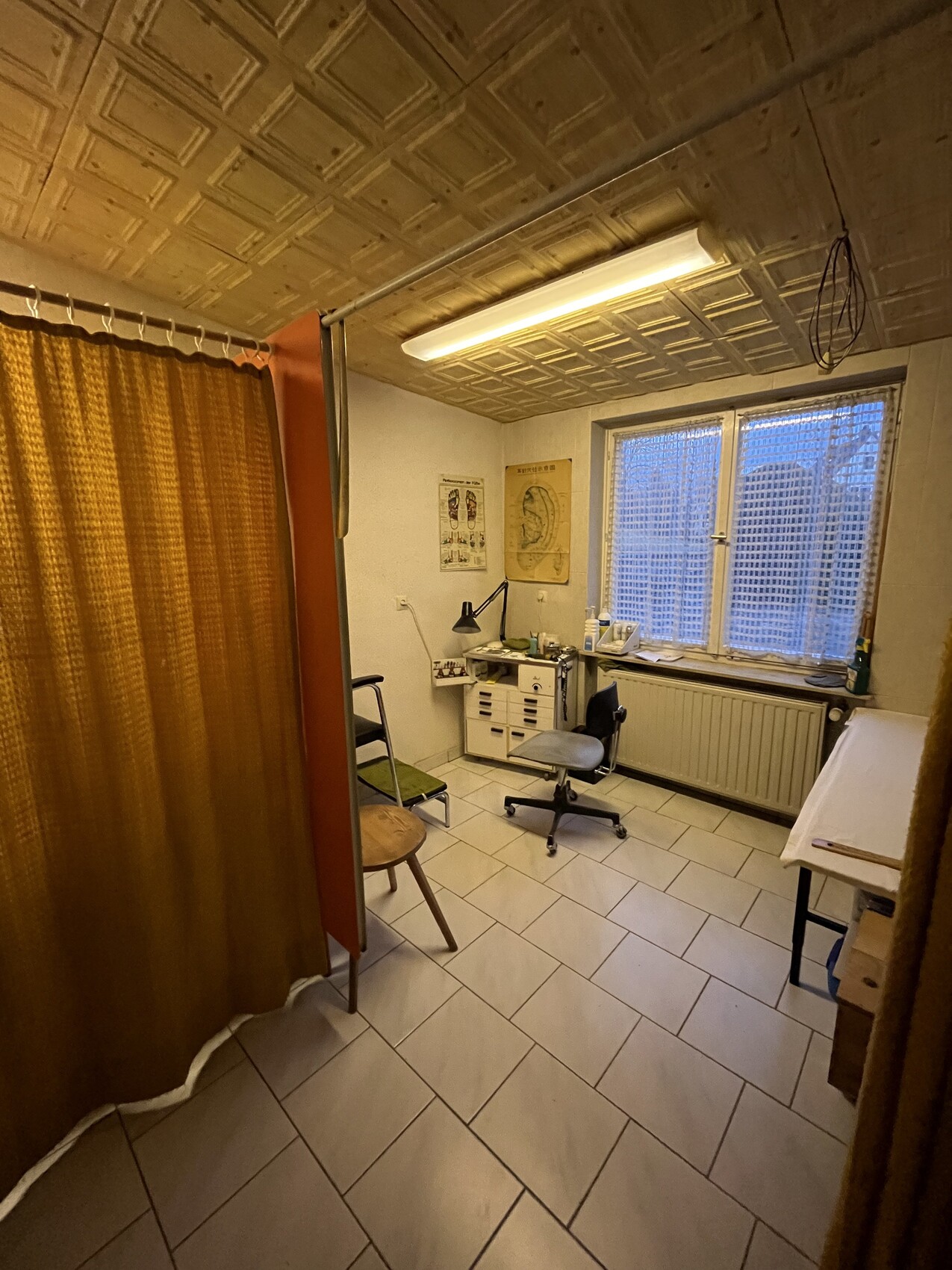 Behandlungsraum mit Vorhang, Schreibtisch, Rollhocker, Poster, Fenster und Untersuchungstisch.