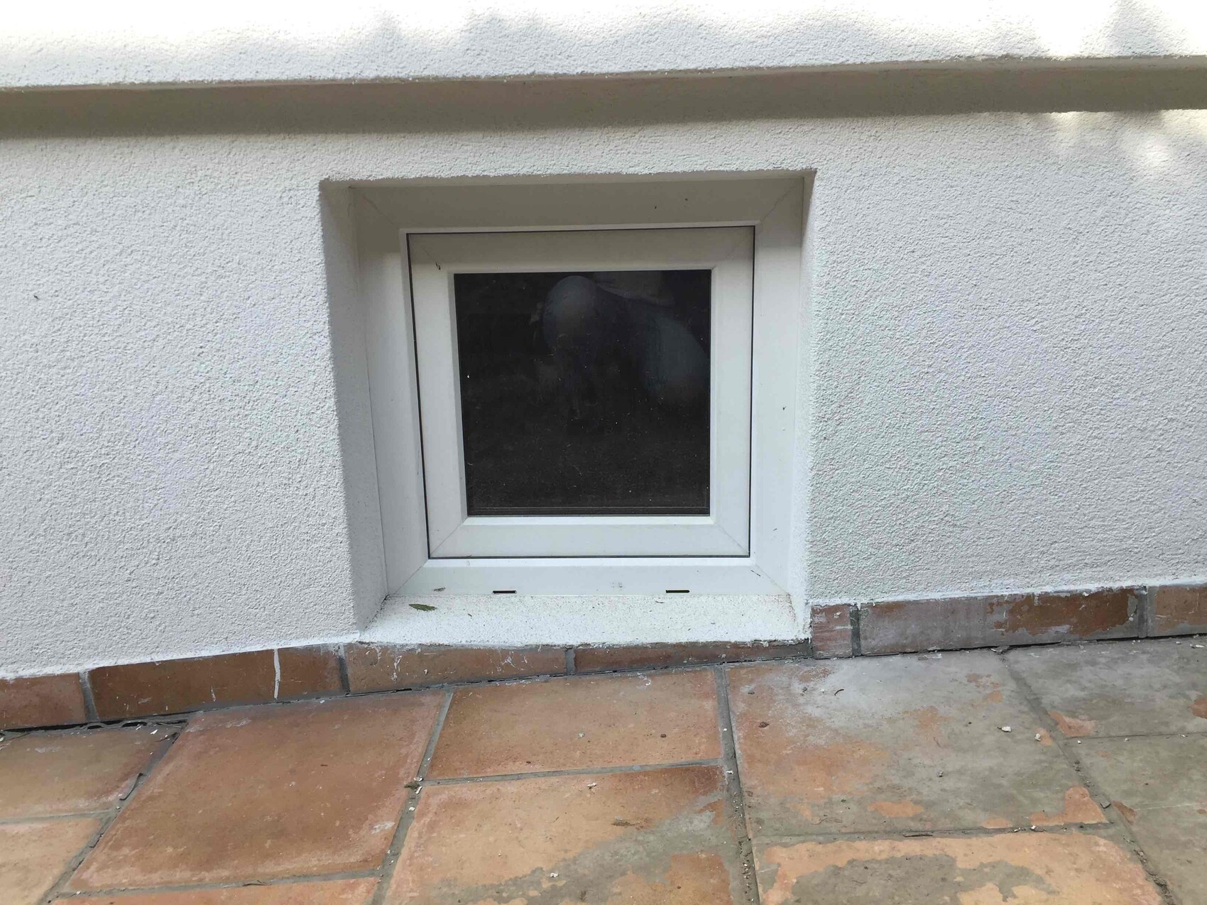 Kleines quadratisches Kellerfenster in weißer Außenwand mit Terrakottafliesen davor