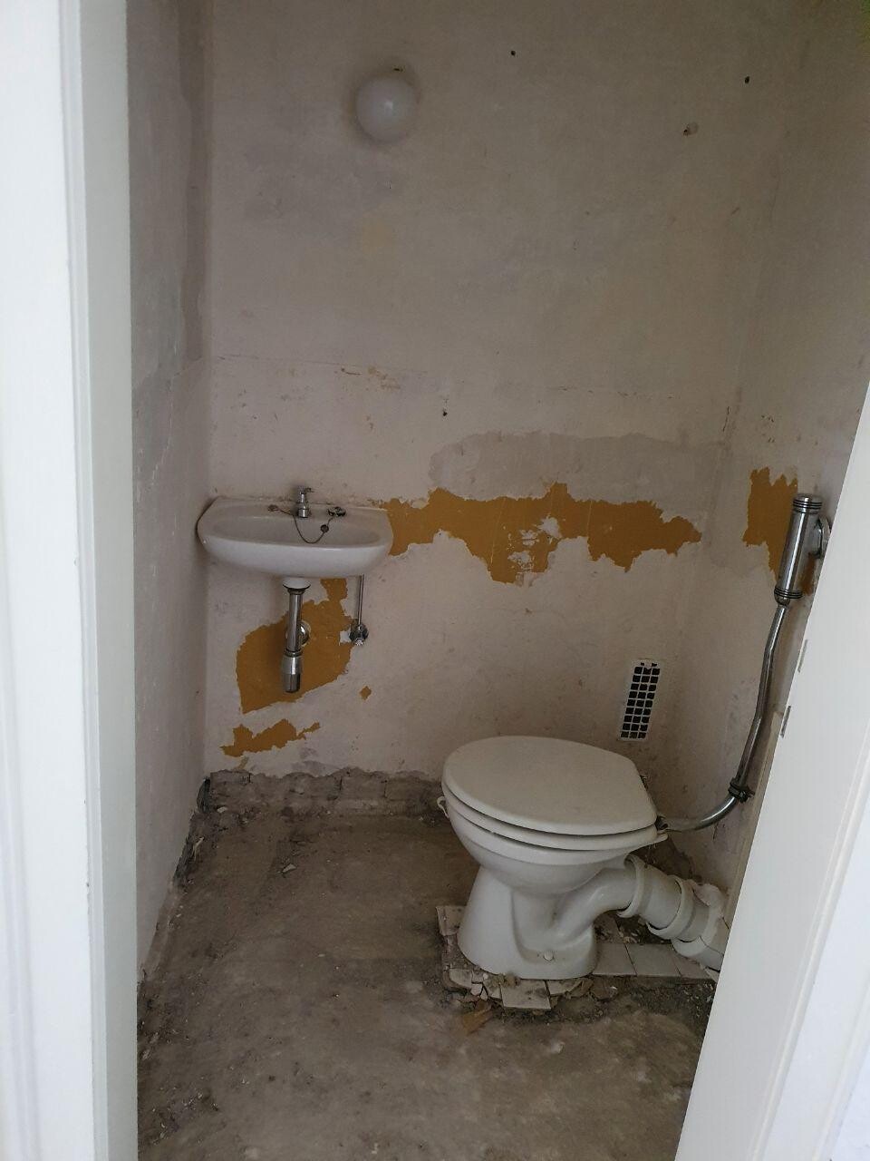 Kleines WC mit abblätternder gelber Wandfarbe, Waschbecken und Toilette, Rohre sichtbar.
