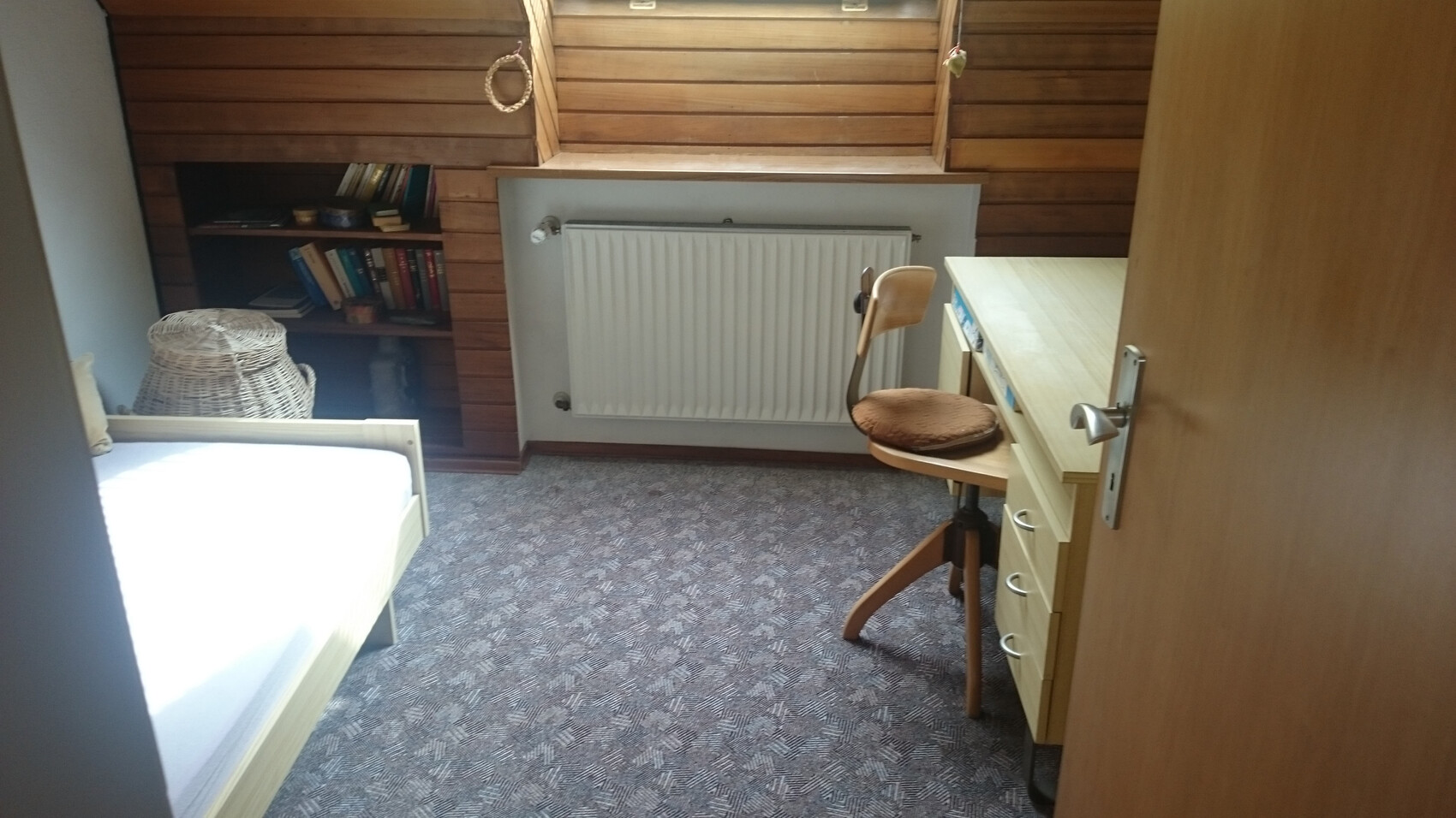Kleines Schlafzimmer mit Holzverkleidung, Bett, Schreibtisch und Regal, Teppichboden.
