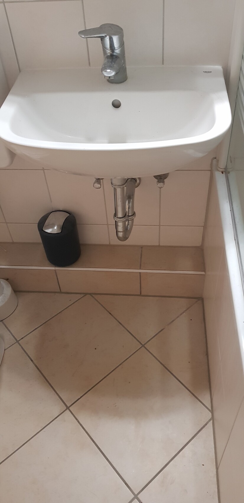 Waschbecken mit Chromarmatur, sichtbare Abflussrohre und schwarzer Abfalleimer auf Fliesenboden.