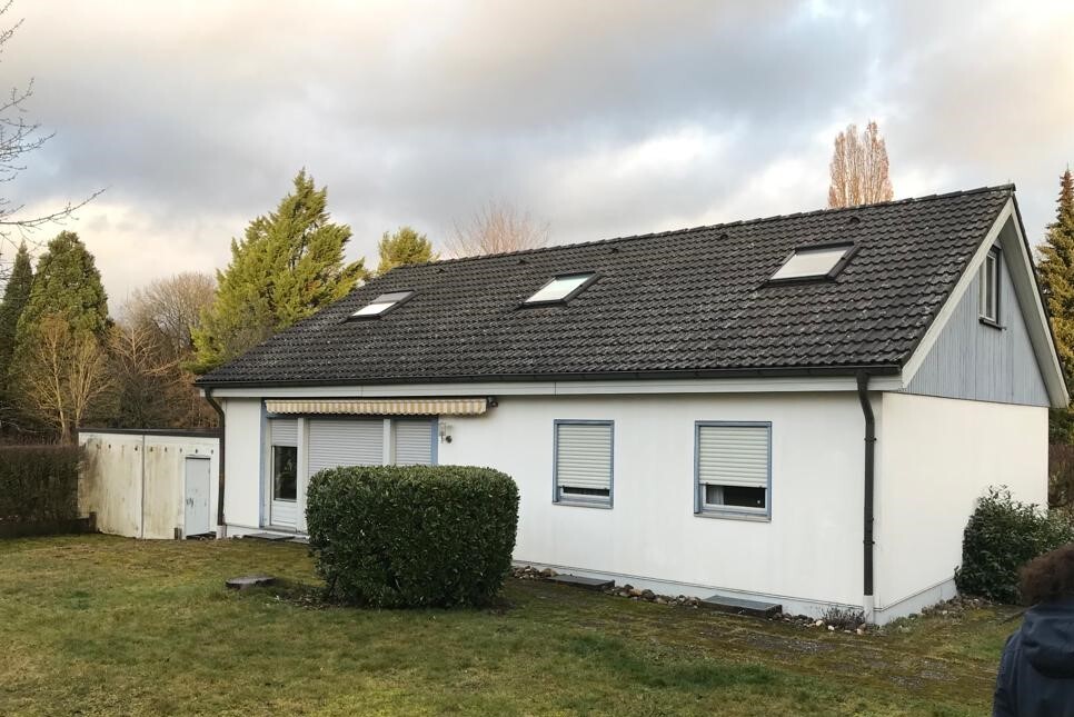 Weißes Einfamilienhaus mit grauem Satteldach und Dachfenstern, Hecke vor Eingang, Schuppen links.
