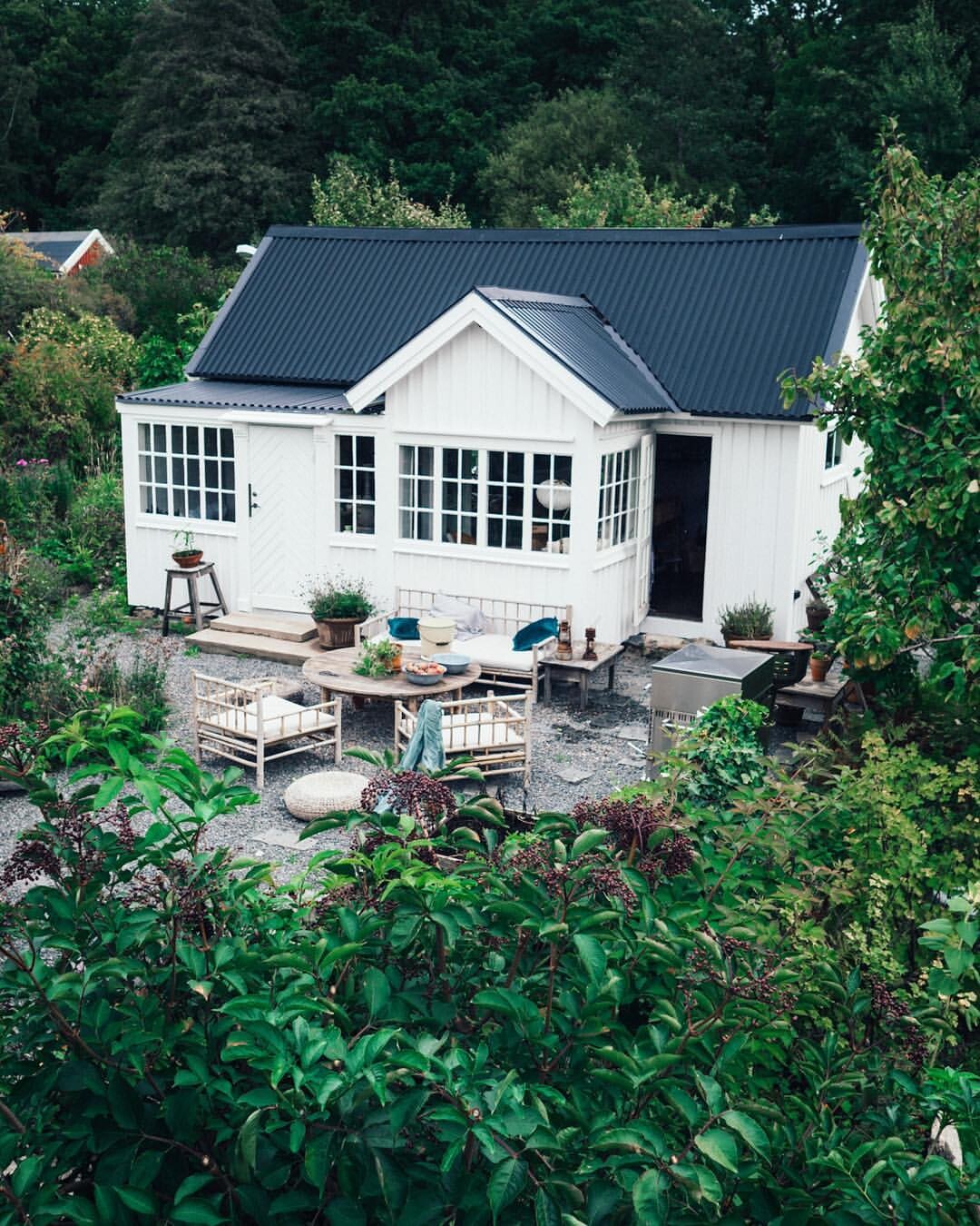 Kleines weißes Gartenhaus mit dunklem Dach, umliegendem Garten und Sitzbereich.