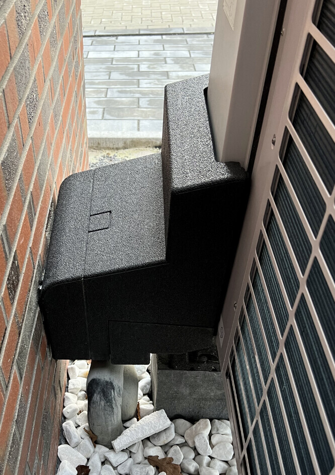Schwarzes Außengerät in engen Spalt zwischen Ziegelwand und Hausfassade auf Kieselsteinen.