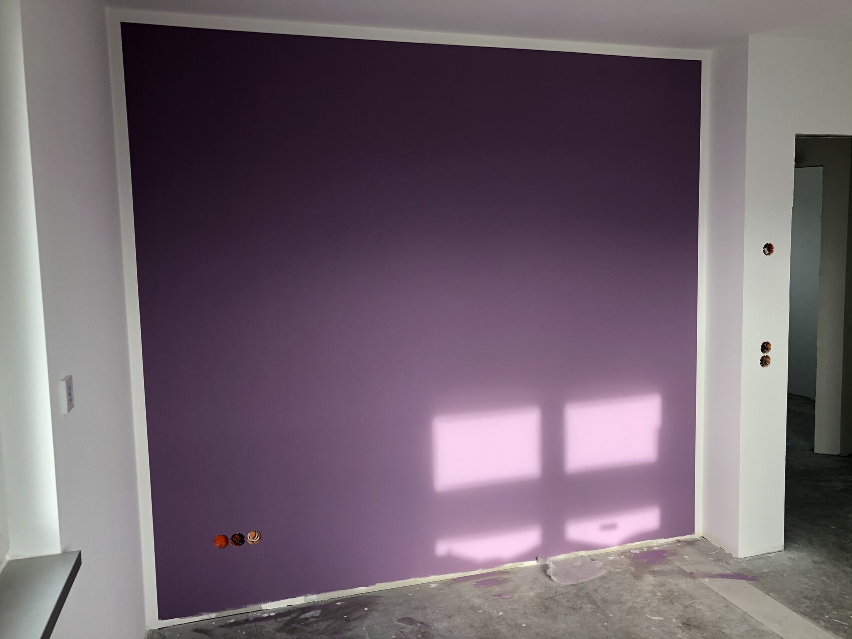Dunkellila Akzentwand zwischen weißen Wänden; Fensterlicht erzeugt rechteckige Lichtflächen im Raum.