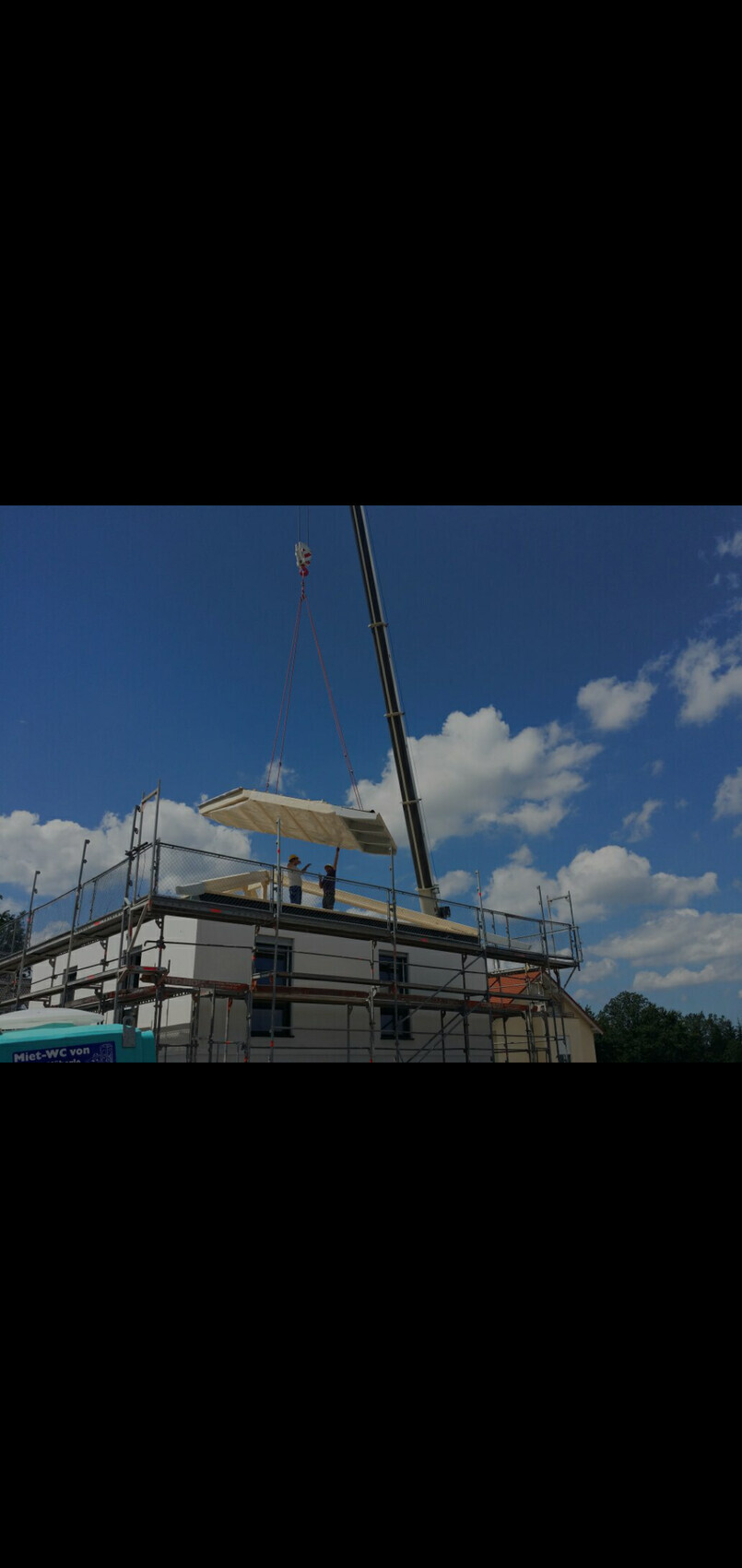 Kran hebt eine große Dachplatte auf ein Baugerüst; zwei Arbeiter sichern das Bauteil.