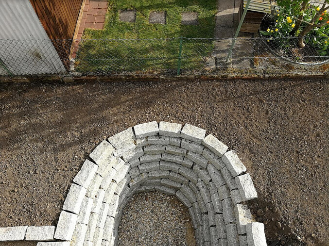 Kreisförmige Mauer aus hellgrauen Steinquadern liegt auf Kiesboden, Garten im Hintergrund.