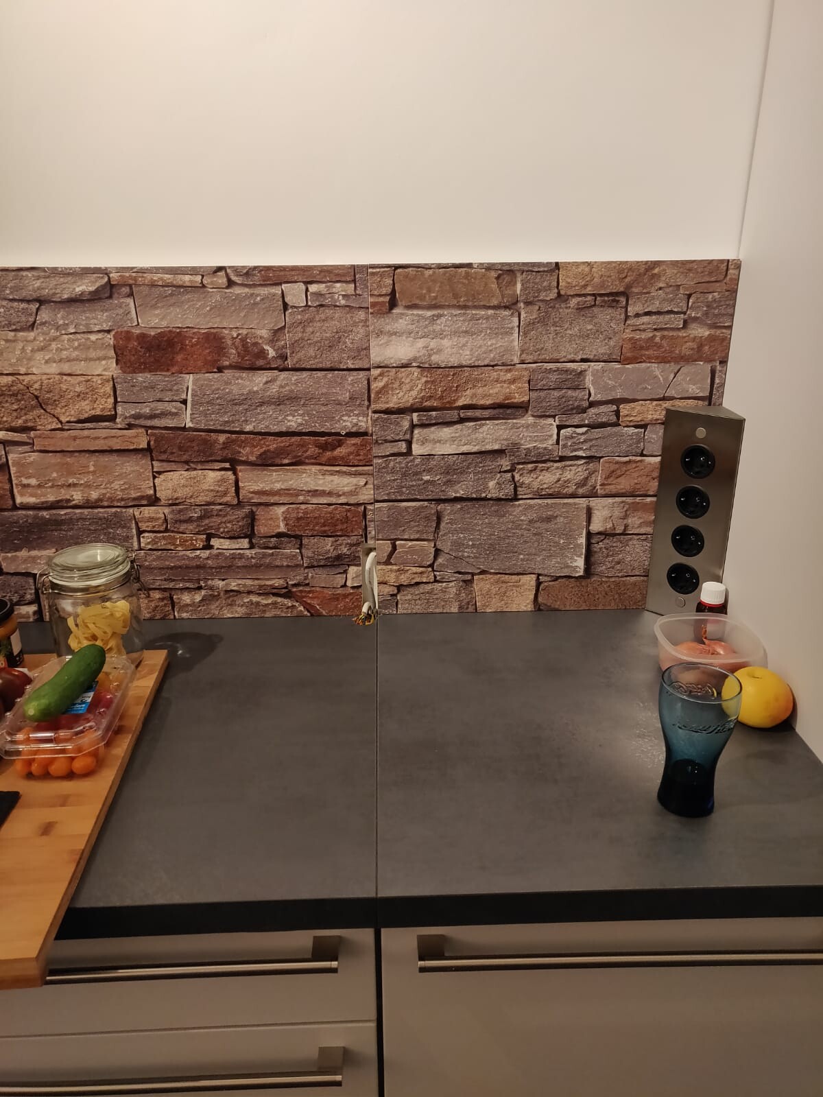 Moderne Küche mit grauer Arbeitsplatte und Steinoptik-Rückwand, Obst auf dem Tisch.