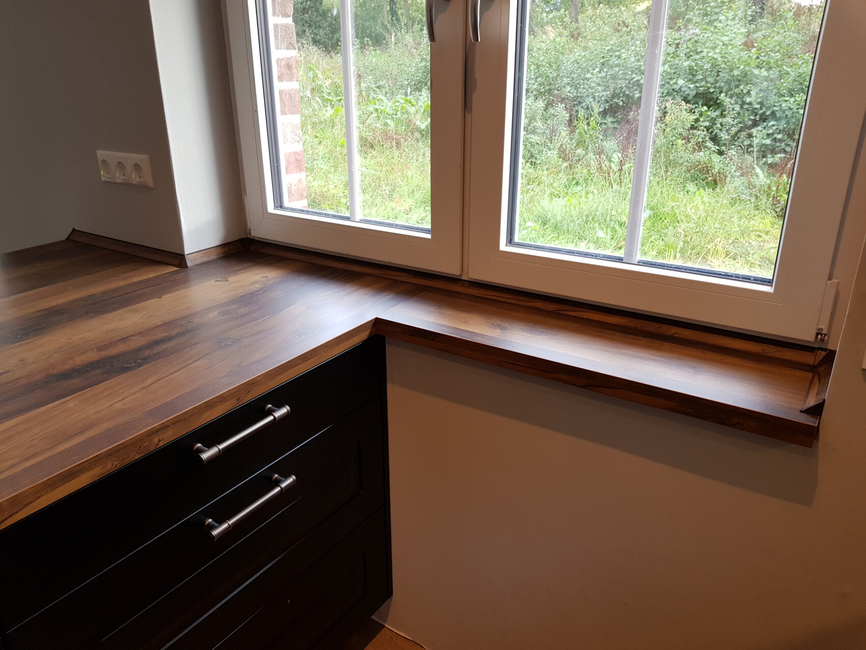 Holz-Arbeitsplatte am Fenster, darunter schwarze Küchenschränke; Blick nach außen in Garten.