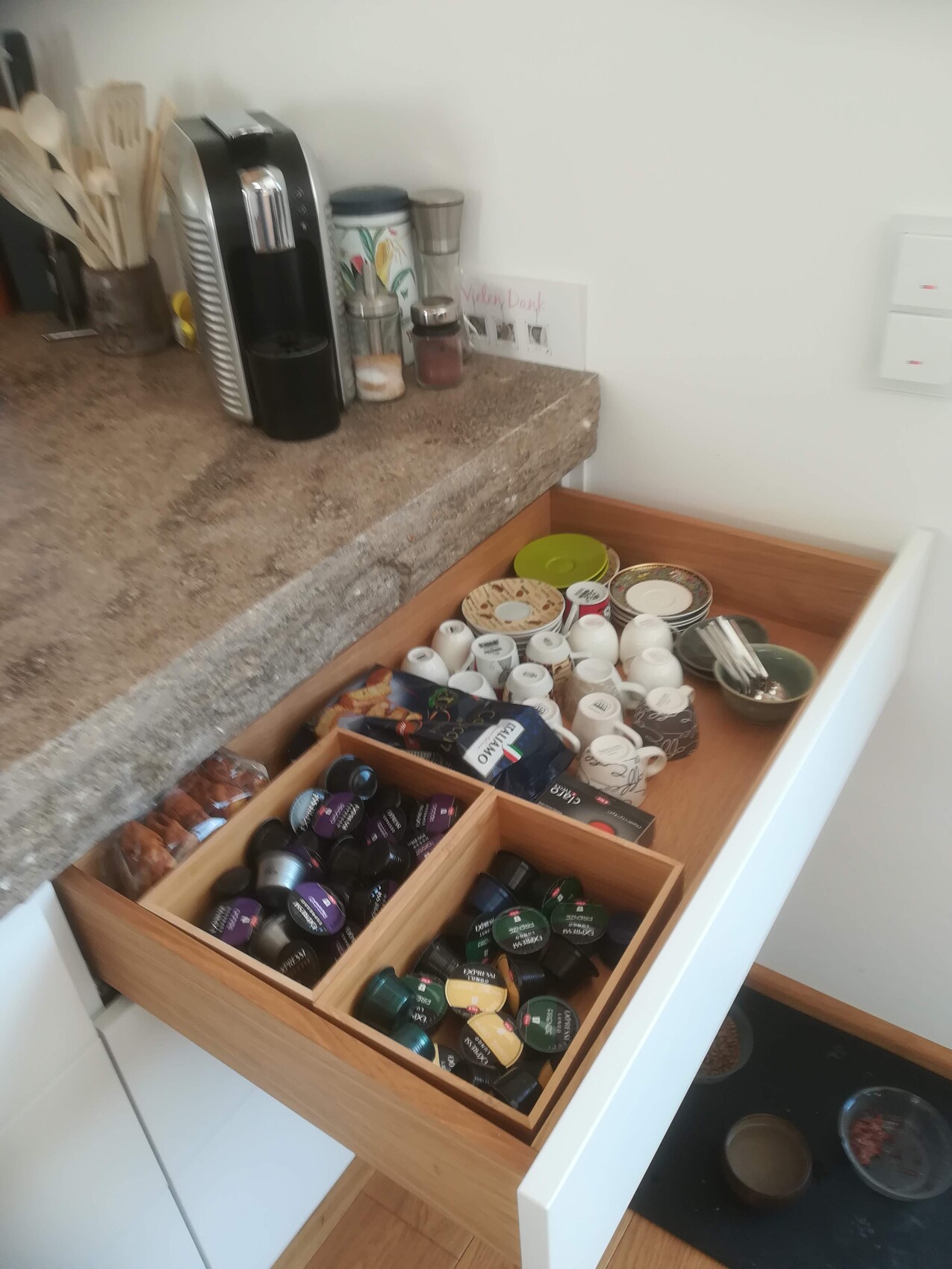 Offene Küchenschublade mit farbigen Kaffeekapseln in Holzfächern, daneben Tassen und Kaffeemaschine.