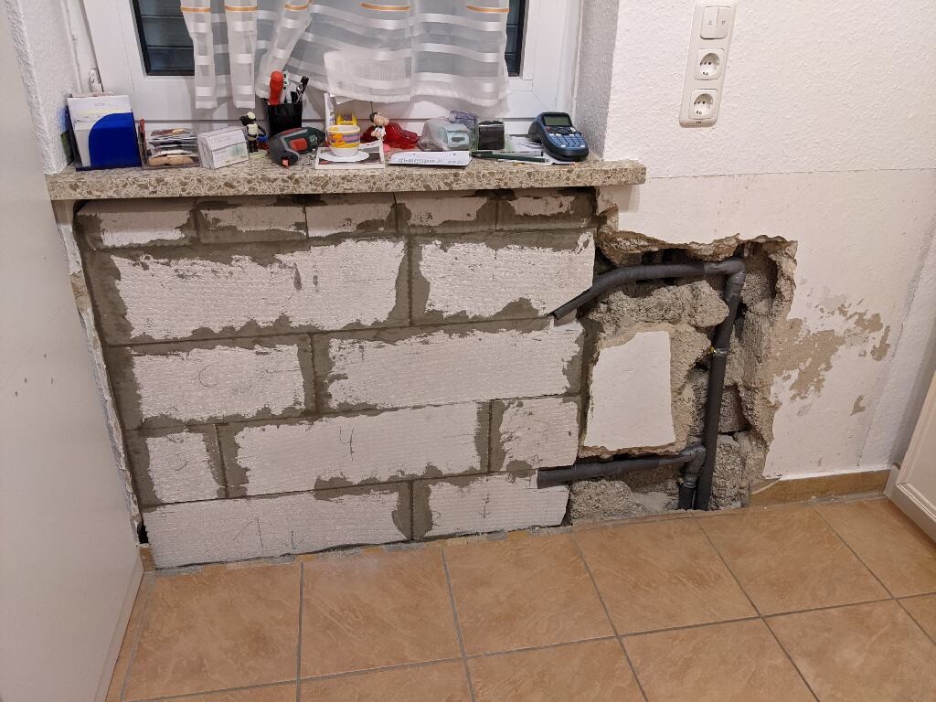 Küche mit freigelegter Betonblockwand, Rohrleitungen im Wanddurchbruch, Werkzeuge auf Fensterbank.