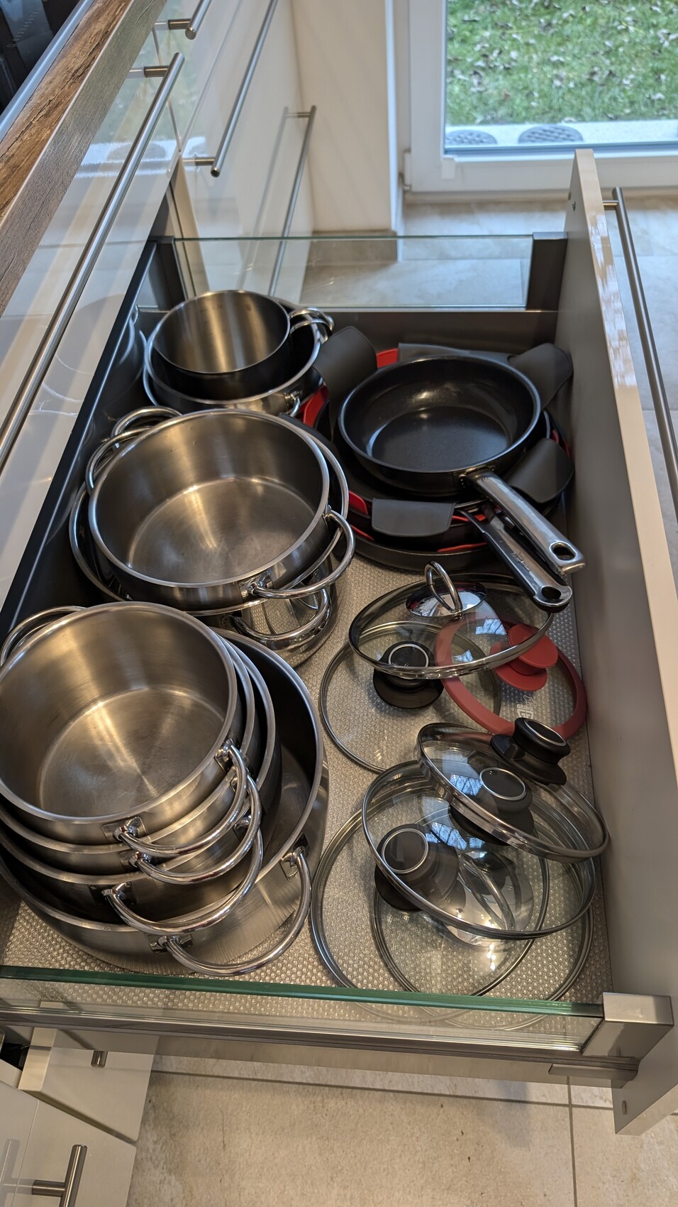 Offene Küchenschublade mit Edelstahl-Töpfen, Pfannen und Glasdeckeln.