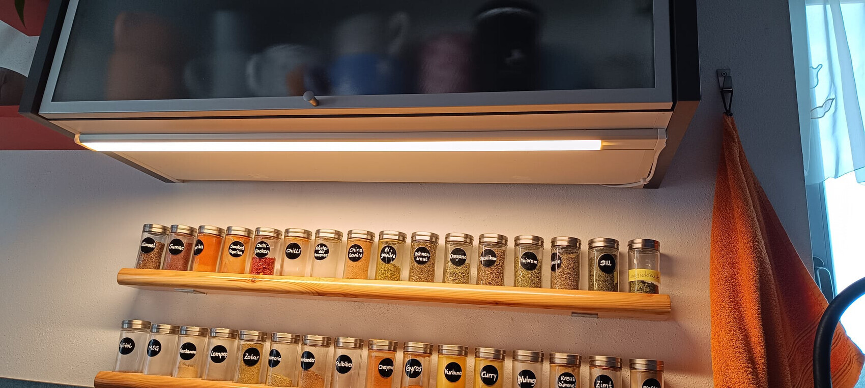 Küche: Glas-Gewürzbehälter auf Holzregalen unter Dunstabzugshaube, orangefarbenes Handtuch rechts.