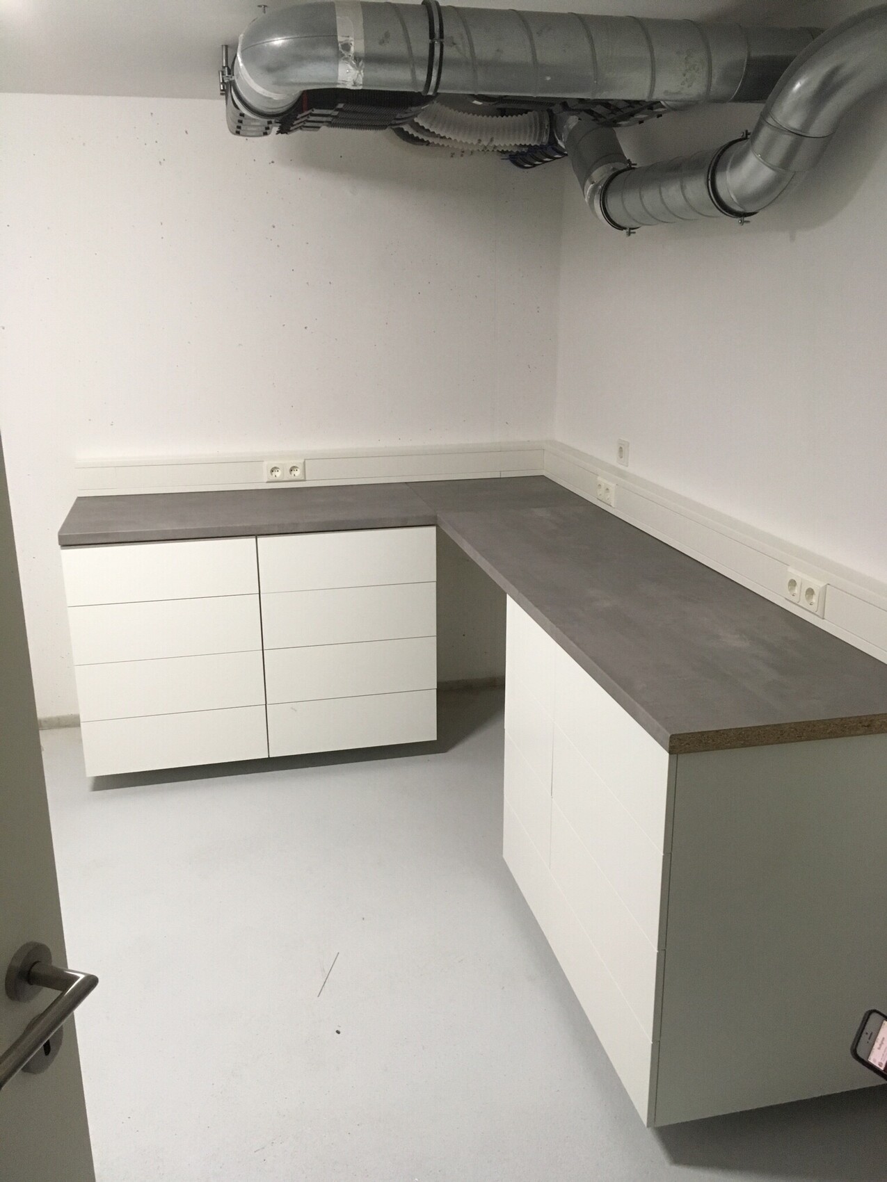 Keller-Küchenbereich mit weißen Schränken, grauer Arbeitsplatte und sichtbaren Lüftungsrohren.
