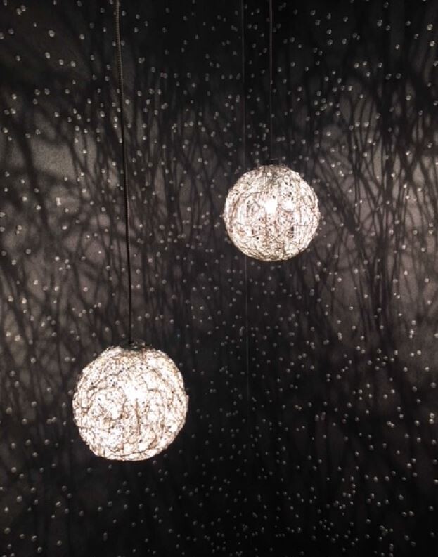 Zwei leuchtende Kugelleuchten hängen an dünnen Kabeln vor dunklem Hintergrund mit vielen Lichtpunkten.