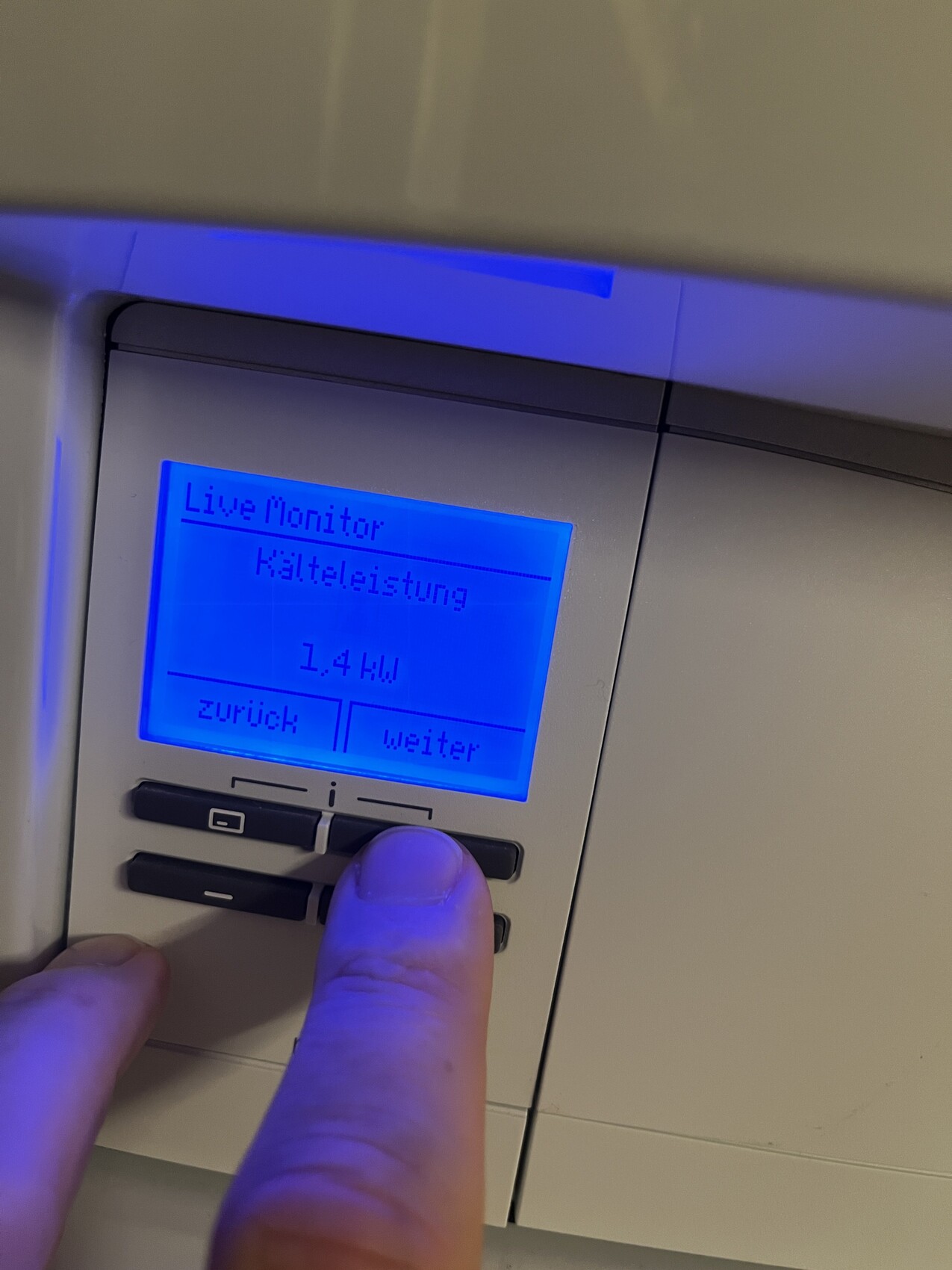 Bedienfeld mit blauem LCD: Live Monitor, Kälteleistung 1,4 kW, Zurück/Weiter; Finger drückt Taste.