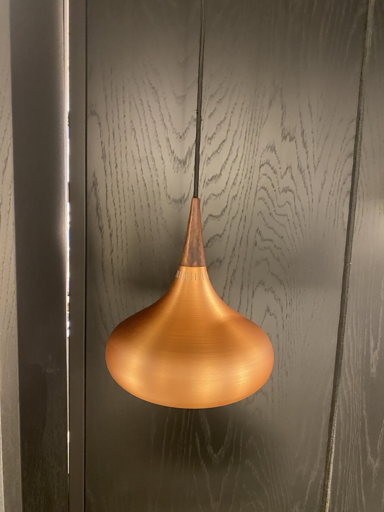 Kupferfarbene Pendelleuchte mit tropfenförmigem Schirm vor dunkler Holzwand.