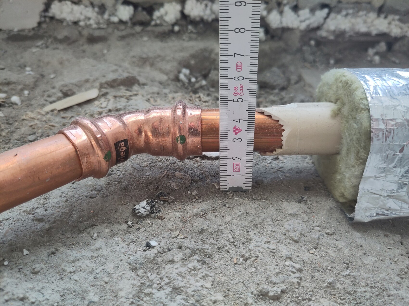 Kupferrohr mit Fitting, daneben Maßband; isoliertes Rohr durch Wandöffnung auf einer Baustelle.